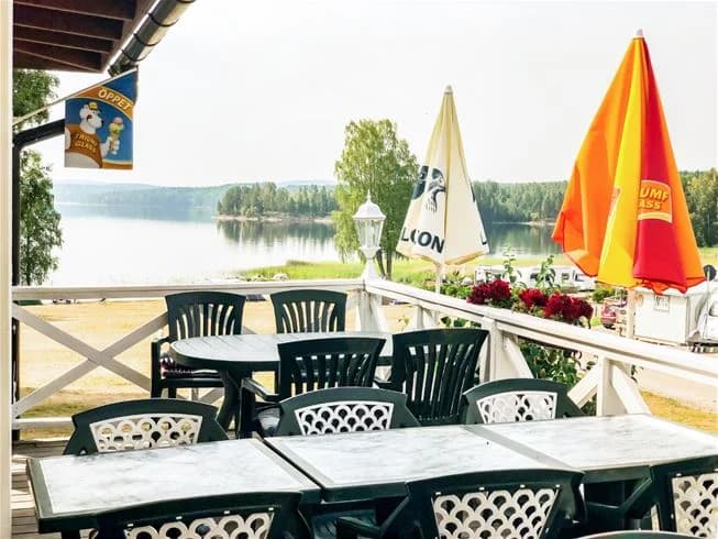 En campingområde där flera trädgårdsmöbler står uppställda utomhus under parasoller. Det finns bord med stolar, och miljön liknar en trevlig utomhusrestaurang. I bakgrunden syns grönska från campingens omgivningar.