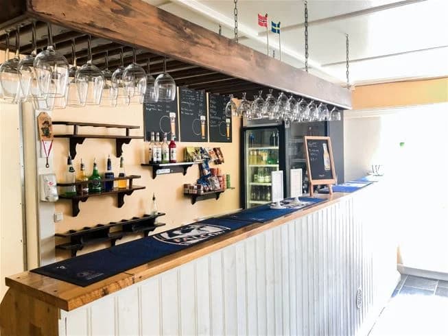 Interiör av en campingbar med hyllor fyllda med flaskor i ett rustikt trädesign. Trätak och bardisk av mörkt trä skapar en varm och inbjudande atmosfär på Charlottenberg camping & stugor - Haganäset.