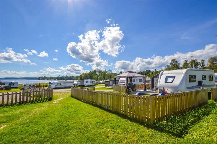 En panoramabild av Charlottenberg camping & stugor - Haganäset, med en lugn reservoar omgiven av grönskande natur. Flera husbilar och husvagnar står parkerade i området, medan fluffiga cumulusmoln svävar över himlen. En idyllisk campingplats som kombinerar natur med komfort.