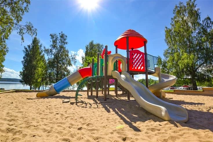 En bild som visar Charlottenberg camping & stugor - Haganäset med lekutrustning, utemöbler, en lekplats och en pool, omgiven av grönska och träd som ger skugga.