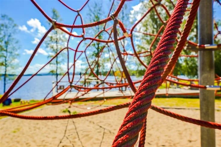 Lekplats utomhus på Charlottenberg camping & stugor - Haganäset, med sandmark och klätterställning i nät, omgiven av grönska i parkliknande miljö.