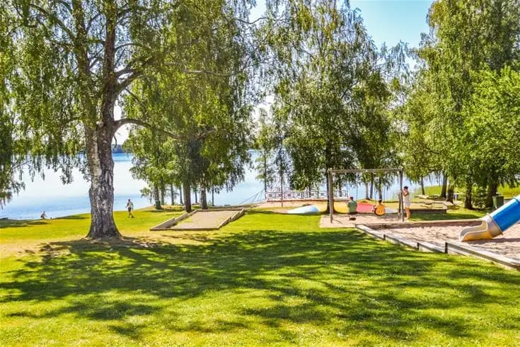 En grönskande campingplats med rymliga gräsmattor och träd som ger skugga, omgiven av ett naturskönt landskap och nära en sjö.