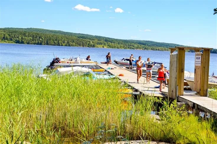 En sommardag vid Charlottenberg camping & stugor - Haganäset med utsikt över en sjö. Ett par båtar ligger vid en brygga, omgivet av grönska och strand. Perfekt för avkoppling och fritidsaktiviteter i en naturskön miljö.