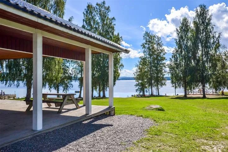 En grönskande campingplats med stora gräsytor, skuggande träd och flera små trädgårdshus. I bakgrunden syns en paviljong och ett glittrande vattendrag.