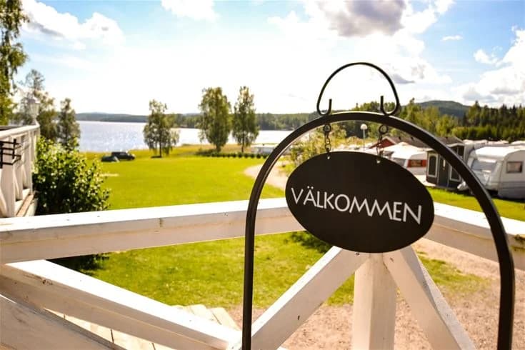 En grönskande campingplats med stora gräsmattor och täta träd, omgiven av lantlig miljö. I bakgrunden syns ett vitmålat hus med rött tak, typiskt för svenska landsbygden.