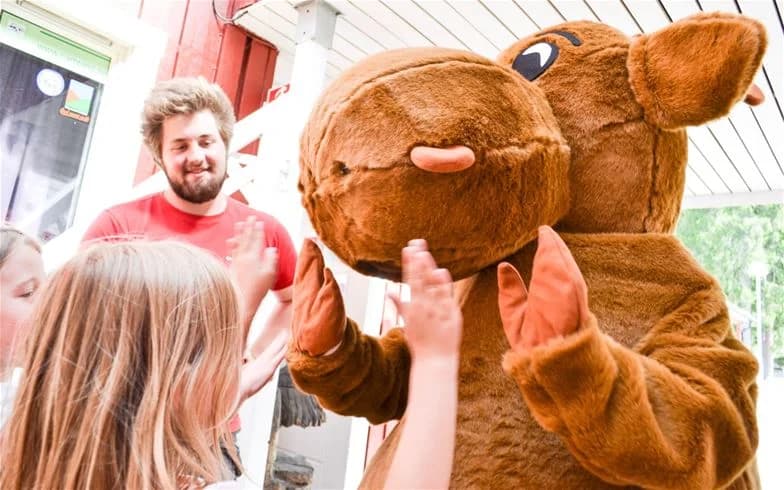 En glad familj poserar framför en leende maskot vid Charlottenberg camping & stugor - Haganäset, vilket framhäver semesterkänsla och glädje.