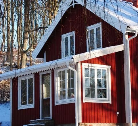 En röd trästuga med vita knutar och sadeltak, omgiven av frodig grönska. Dörren är vit med glasruta och flera spröjsade fönster pryder väggarna.