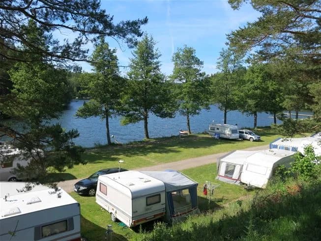 Bilden visar ett naturskönt område vid Yxningens Camping med utsikt över en sjö. I förgrunden syns en grön gräsmatta och en liten stig som leder ner till sjöns strandkant. Området omges av lummiga träd och det stilla vattnet reflekterar den blå himlen.