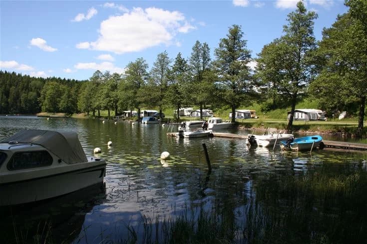 En liten båt ligger förtöjd vid stranden av en sjö omgiven av lummiga träd, med spegelblankt vatten som speglar den blå himlen.