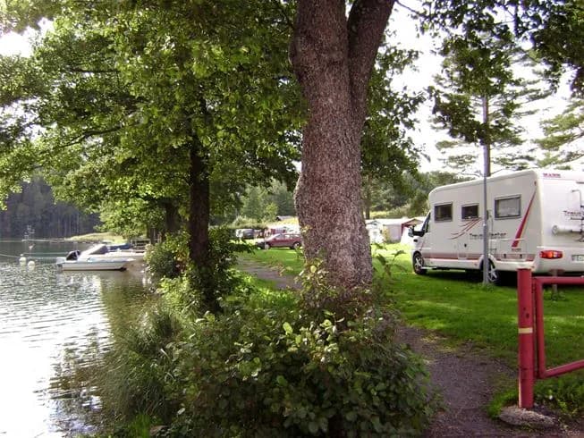 Bild av en naturskön camping vid sjön Yxningen. Träd omger platsen och flera båtar ligger förtöjda vid vattnet. En husvagn och andra campingfordon står parkerade i området, vilket antyder en populär rekreationsplats nära naturen.