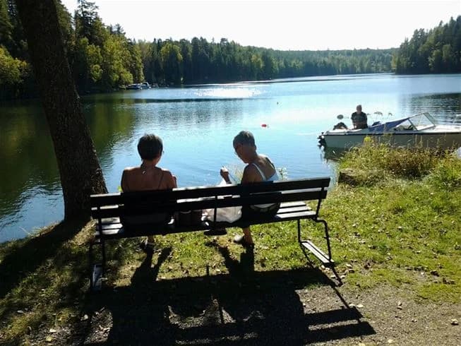 En somrig scen vid Yxningens camping, där besökare njuter av rekreation vid sjön. Några båtar syns vid strandkanten, och människor ägnar sig åt olika fritidsaktiviteter i det natursköna området.