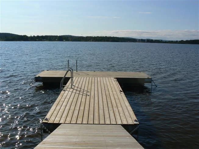 Alt-text: En träbrygga sträcker sig ut över en stilla sjö vid Storstrand kursgård, omgiven av lummig grönska och blå himmel.
