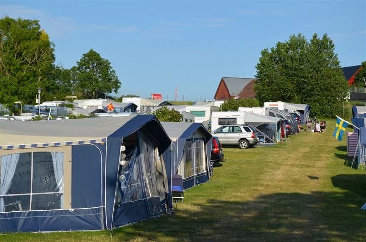 En bild av Klinta Camping som visar tält, parkerade husvagnar och husbilar, campingstolar samt en presenning som ger skugga. I bakgrunden syns en parkering för besökare.