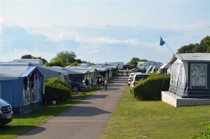 En grön campingplats med flera husbilar parkerade. Besökare i somriga badkläder rör sig mellan fordonen, vilket skapar en avslappnad semesteratmosfär.