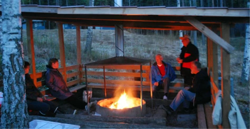 En mysig lägereld vid trästolar och en timmerstuga på Sölje Camping, omgiven av skog och rustik campingmiljö.