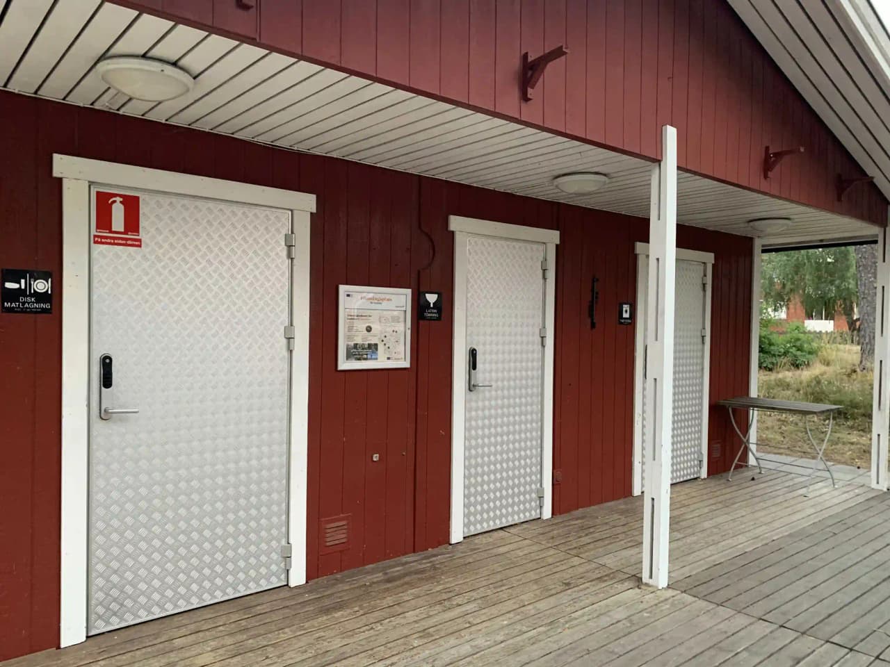 En träfasad med en dörr visas, troligen en stuga eller husvagn vid Joelskogens camping & ställplats. Fasadens trä är behandlat med en träbets som framhäver materialets naturliga ådring.