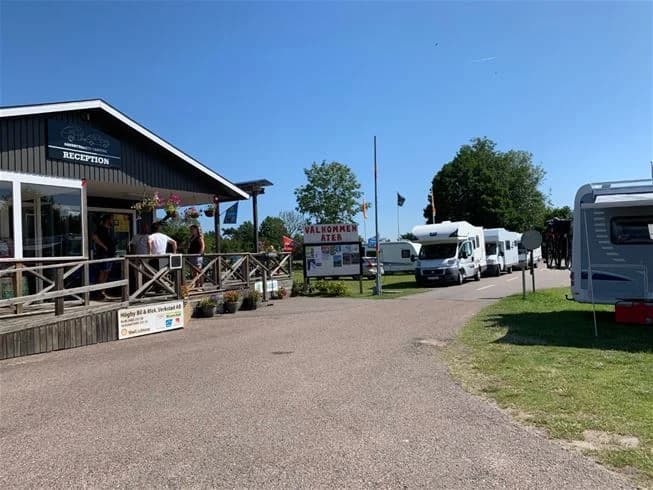 En solig campingplats vid Sandbybadets camping med parkerade fordon, inklusive husbilar, skåpbilar och en SUV, samt uppställda tält. Campingdeltagare njuter av en avslappnad atmosfär på den rymliga parkeringen.