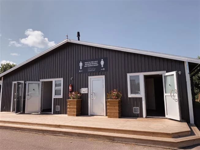 Bild av en träbyggnad vid Sandbybadets camping, med träpaneler och en enkel dörr, omgiven av grönska och trädgårdselement.