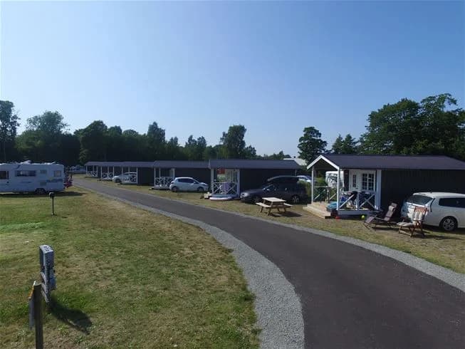 En parkeringsplats vid Sandbybadets camping med flera husbilar och campingfordon uppställda. Lummig omgivning med träd och grönområden i bakgrunden.