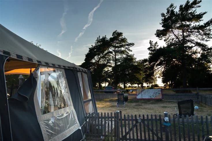 En campingplats med ett uppställt tält under en skuggande presenning. En sportig SUV och en kompakt MPV är parkerade nära tältet. Området ser ut att vara en del av Haga Park Camping & Stugor.
