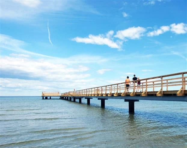 En solig dag vid Köpingbaden Camping, med en lång brygga som sträcker sig ut i det glittrande vattnet. I fjärran syns horisonten smälta samman med den lätt molniga himlen.