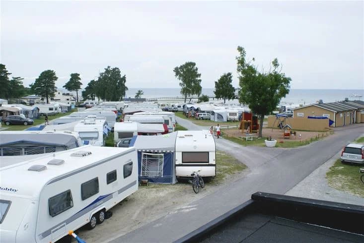 En campingplats med flera husvagnar och husbilar parkerade på grusade områden. En stor lastbil syns i bakgrunden. Det är en solig dag med några träd i närheten.