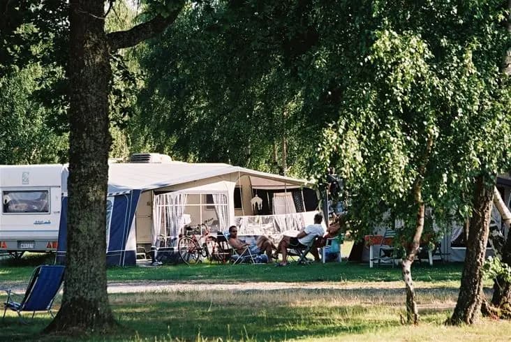 En bild av Saxnäs camping visar tält och husvagnar uppställda på en grön gräsmatta, omgivna av höga träd som ger skugga. Utomhusmöbler syns i förgrunden, och besökare njuter av fritid i en naturskön miljö.