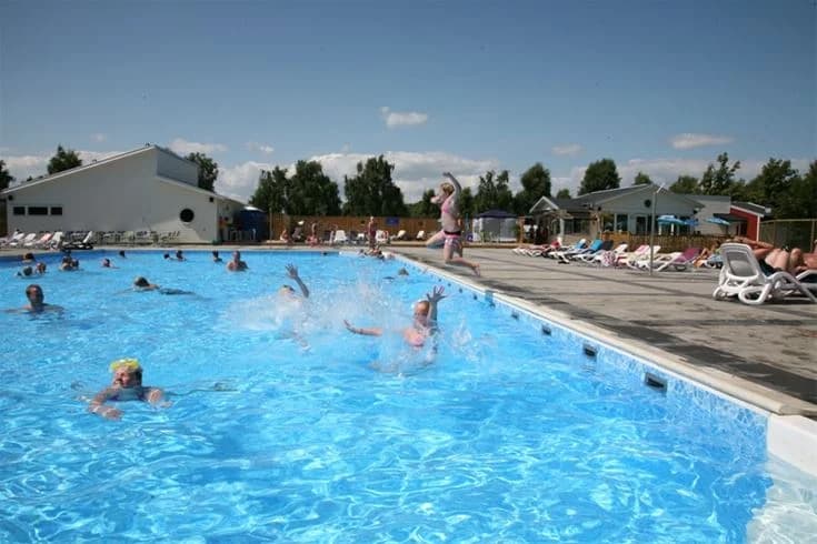 En bild av en utomhusbadplats på Saxnäs Camping, där besökare njuter av semester och fritid. I förgrunden syns personer som badar och solar vid poolområdet. Bakgrunden visar en resortliknande miljö med en avslappnad och lekfull atmosfär.