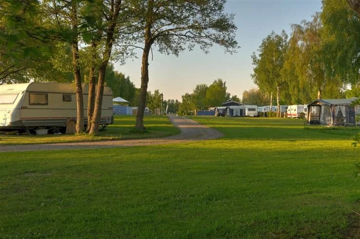 En stor, öppen campingplats vid Saxnäs camping med frodiga gräsmattor och omgivande träd. Ett par husvagnar och en lastbils-semitrailer är parkerade på området.