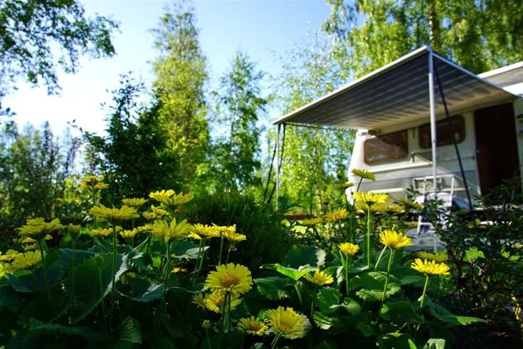 En husvagn står parkerad i en grönskande trädgård med skugga från träd. Det finns utemöbler på gräsmattan, idealiskt för avkoppling vid Otterbergets bad och camping.