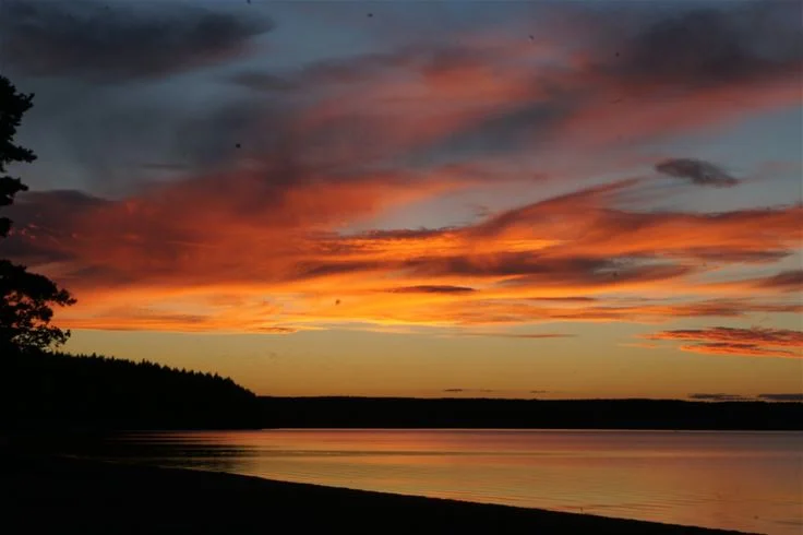 En vacker solnedgång över ett stilla vattendrag vid Otterbergets bad och camping, med mjuka moln på himlen och en horisont som speglas i vattnet.