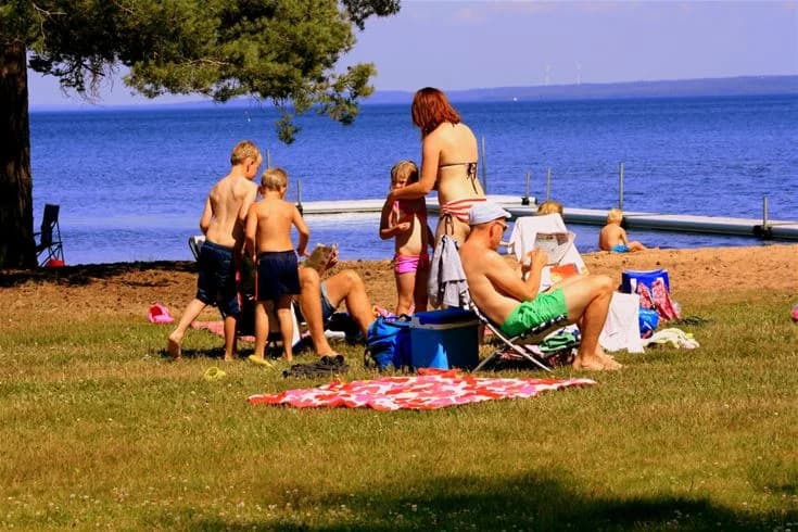 En solig sommardag vid Otterbergets bad och camping, där människor njuter av fritidsaktiviteter vid kusten. Personer kopplar av, solbadar och upplever semesterkänslan nära havet.
