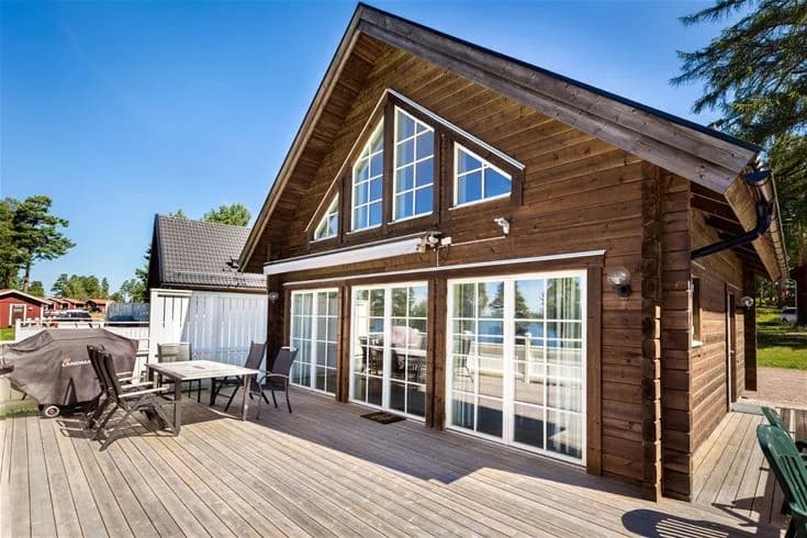 En naturskön vy av en trästuga vid Leksand Resort, komplett med trädäck och en inbjudande veranda. Stugan omges av frodig grönska och är byggd med hållbara trämaterial, vilket ger en varm och mysig atmosfär.