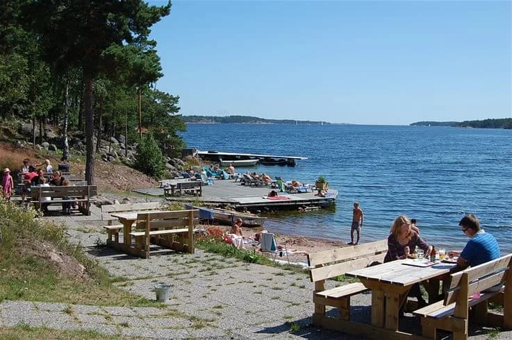 En somrig strandscen vid tättö havsbad & camping med människor som njuter av sol och bad. Utemöbler syns på stranden vid kustlinjen, med det glittrande vattnet i bakgrunden.