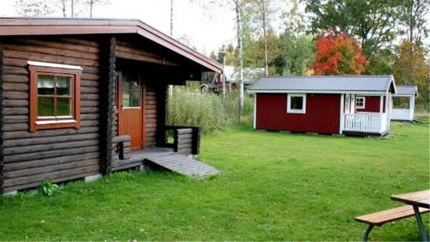 En liten timmerstuga vid Emmaboda camping, omgiven av grönskande träd. Stugan har en enkel träfasad med en dörr och fönster, belägen på en lugn och naturnära plats.