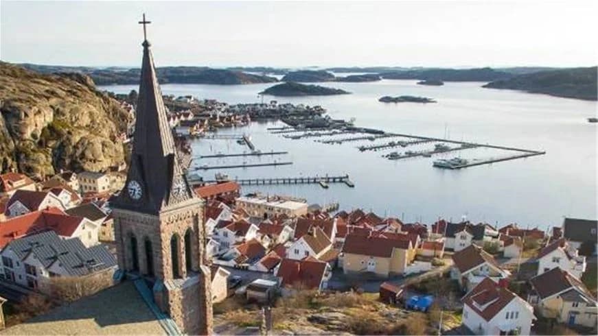 En översikt av Fjällbacka camping med vy över stadens hustak, en kyrkspira synlig i bakgrunden, omgiven av kustområden och en bostadsområde.