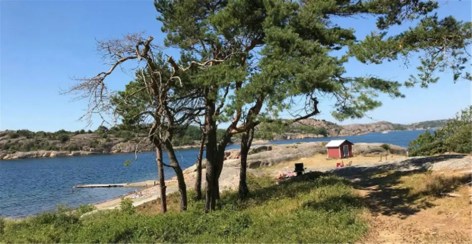Vy över Fjällbacka camping vid kusten, med en sandstrand och sjö i förgrunden samt bergiga landformer och skärgård i bakgrunden.