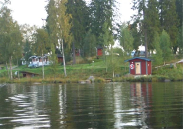 En idyllisk bild av Munkebergs Camping med en pittoresk stuga vid kanten av en sjö, omgiven av frodig grönska och klart vatten.