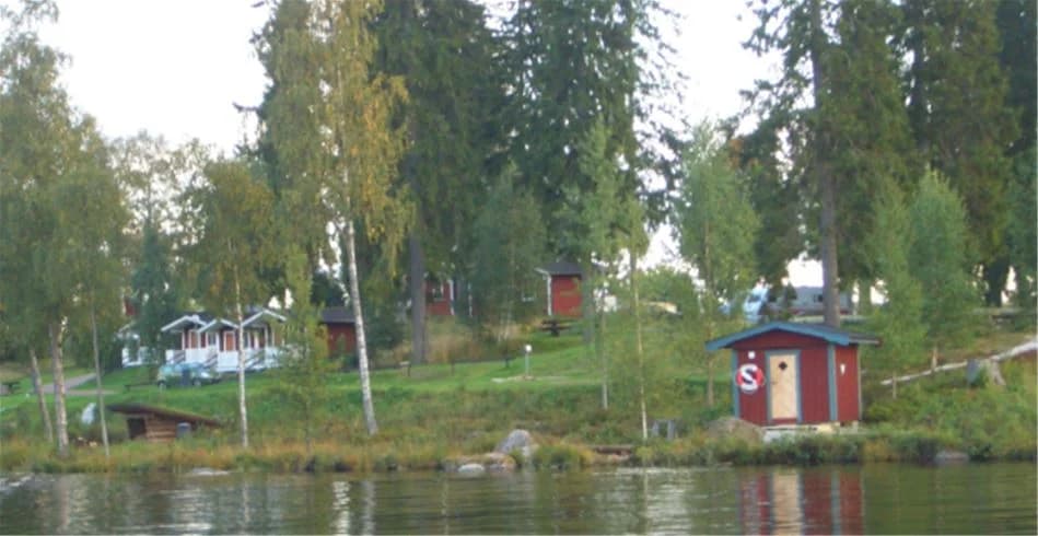 En pittoresk campingplats vid Munkebergs camping med en liten stuga nära kanten av en lugn sjö. Runt sjön finns frodiga träd och växter som bidrar till den natursköna miljön.