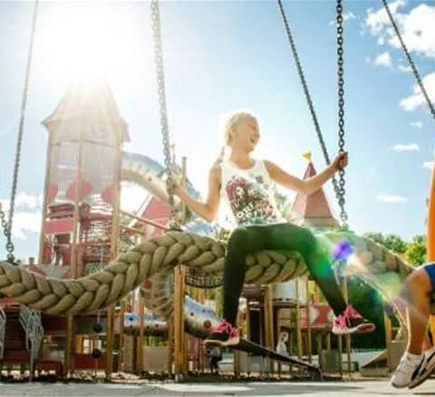En grupp glada människor njuter av en sommardag på Gustavsvik Resort. De leker på en färgglad lekplats med olika utomhuslekredskap. Fötter in sneakers syns i förgrunden, vilket framhäver känslan av fritid och rekreation i en offentlig parkmiljö.
