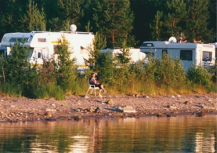 En husvagn står parkerad vid en sjö på Storsands camping, med en strand och skog i bakgrunden. Perfekt för rekreation och naturupplevelser.