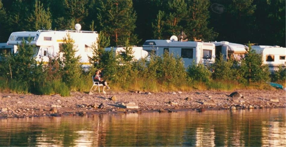 En vidsträckt campingplats vid en sjö med en husbil parkerad nära strandkanten. Vattnet glittrar under en klar himmel medan det omgivande landskapet har frodig grönska. Perfekt för utomhusaktiviteter och naturnära upplevelser.