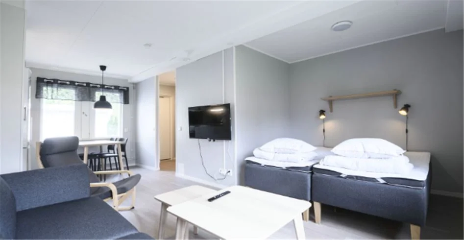 En ljus och rymlig campingstuga med enkel inredning. Rummet innehåller en bekväm säng med vita sängkläder, ett trägolv och ett fönster som släpper in naturligt ljus.