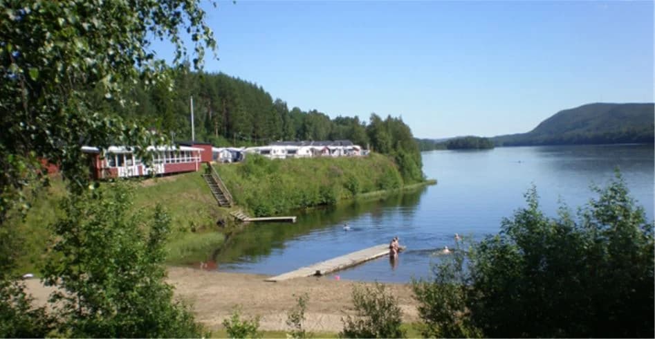 En naturskön vy över Kävsta camping med en stilla sjö omgiven av grönskande träd längs strandkanten.