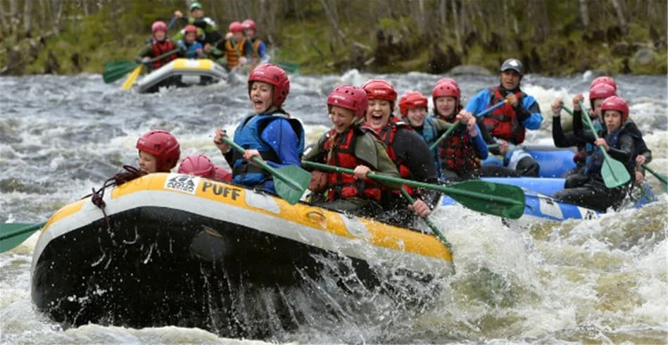 En grupp människor iklädda flytvästar paddlar en uppblåsbar båt nerför en fors omgiven av grönskande natur vid Idre Fjäll Camping.