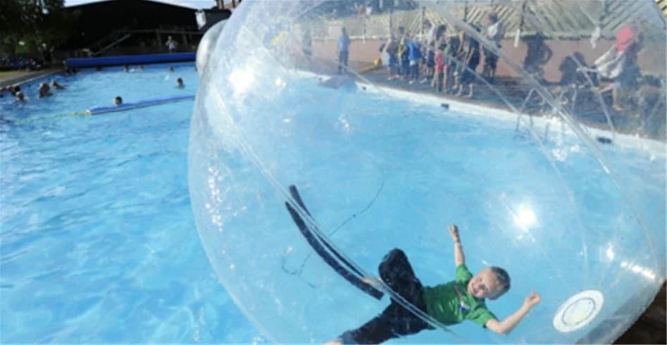 En familj leker i en stor utomhuspool på Idre Fjäll Camping, omgiven av grönskande berg och med uppblåsbara badleksaker som flyter på vattnet.