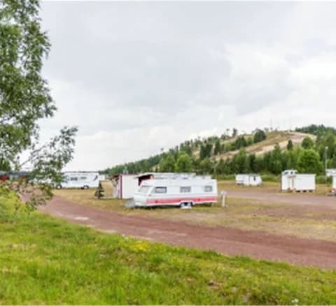 En rad husbilar och husvagnar står parkerade på en öppen yta vid Orsa Grönklitt, omgiven av grönskande natur och blå himmel.