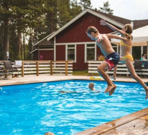 En grupp människor har roligt och simmar i en pool på Orsa Grönklitt, ett rekreationsområde. Några hoppar in i vattnet klädda i badkläder under en solig semesterdag.