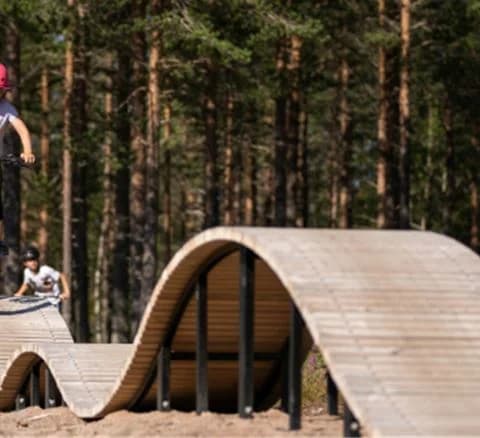 En person cyklar genom en grön skog i Orsa Grönklitt, omgiven av träd. Cyklisten bär en cykelhjälm och kör på en mountainbike, vilket understryker platsens sceniska och äventyrliga miljö.