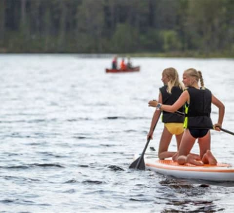 En person står och paddlar på en SUP-bräda mitt på en lugn sjö omgiven av skog. Det är en solig dag, och vattenytan glänser i solljuset.